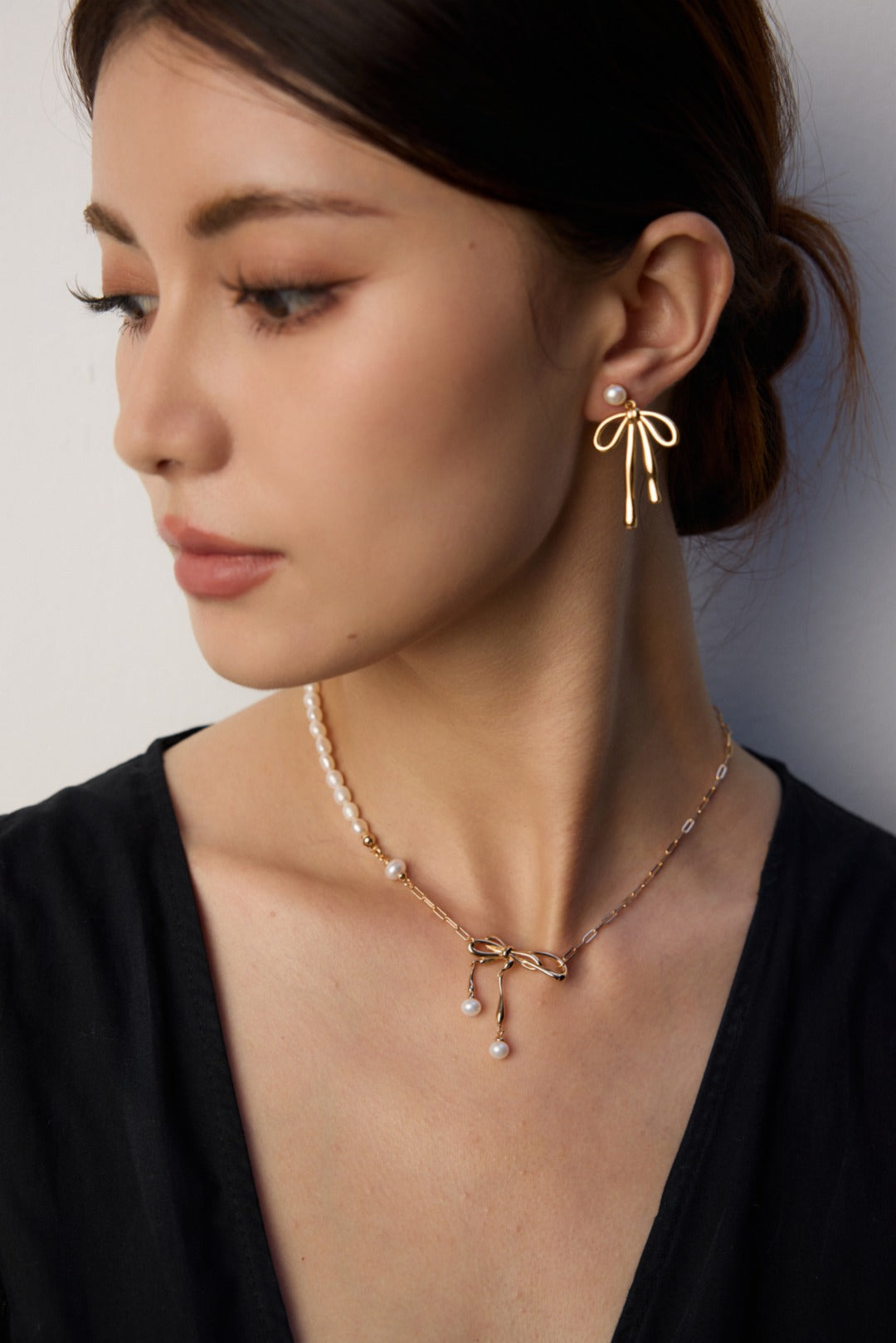 A model wearing a luminous natural freshwater pearl stud earrings with a dangling high-polished fluid gold metallic ribbon bow knot pendant