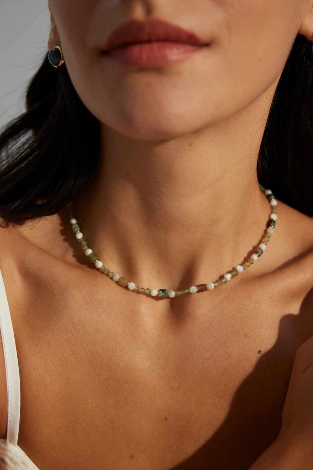 A model wearing an art-gallery style beaded strand necklace featuring multi-shaped natural green moss agates, faceted micro beads, and earth-tone stone cylinders