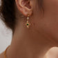 Close-up of a person wearing gold earrings with green gemstones against a neutral background