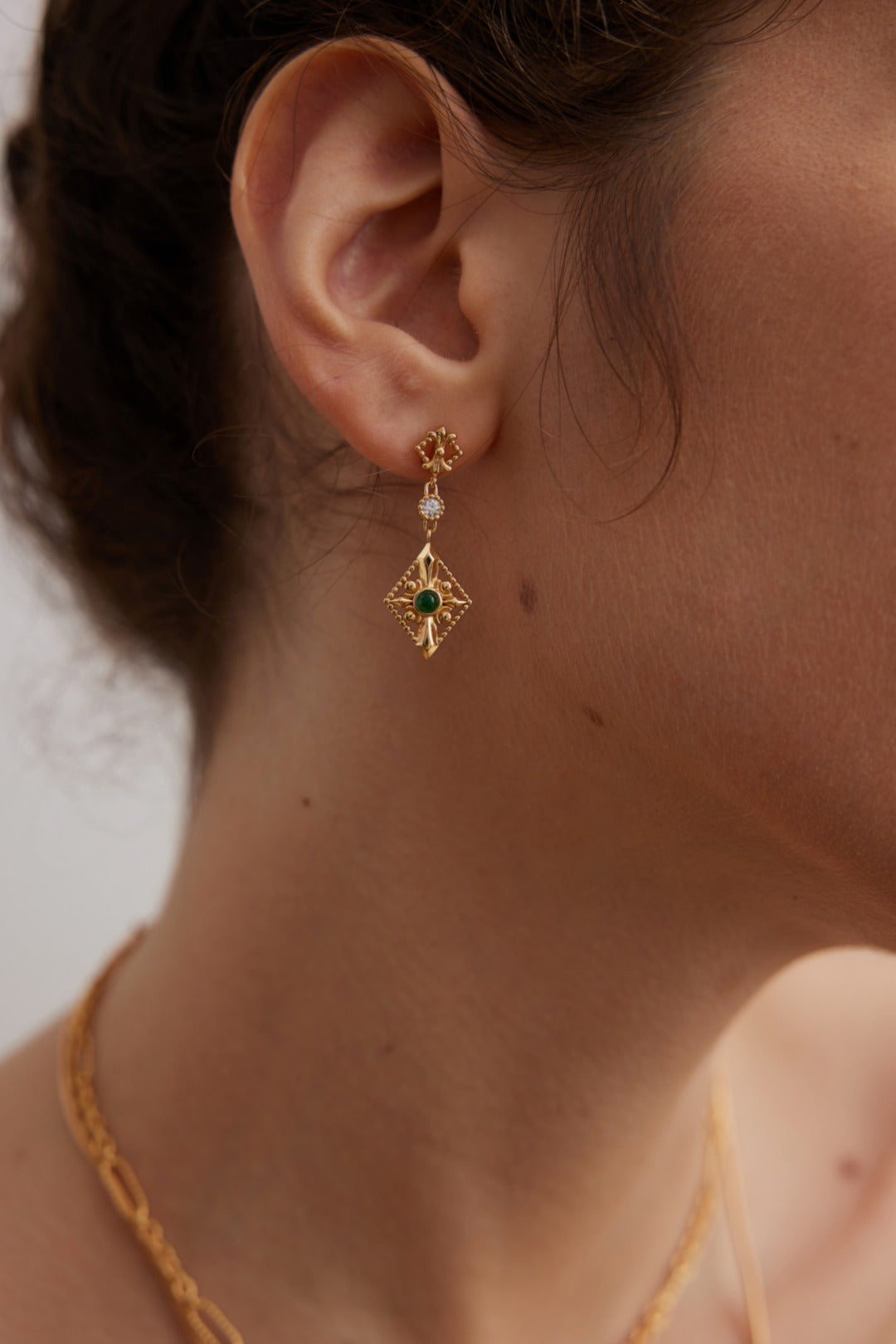 Close-up of a person wearing gold earrings with green gemstones against a neutral background